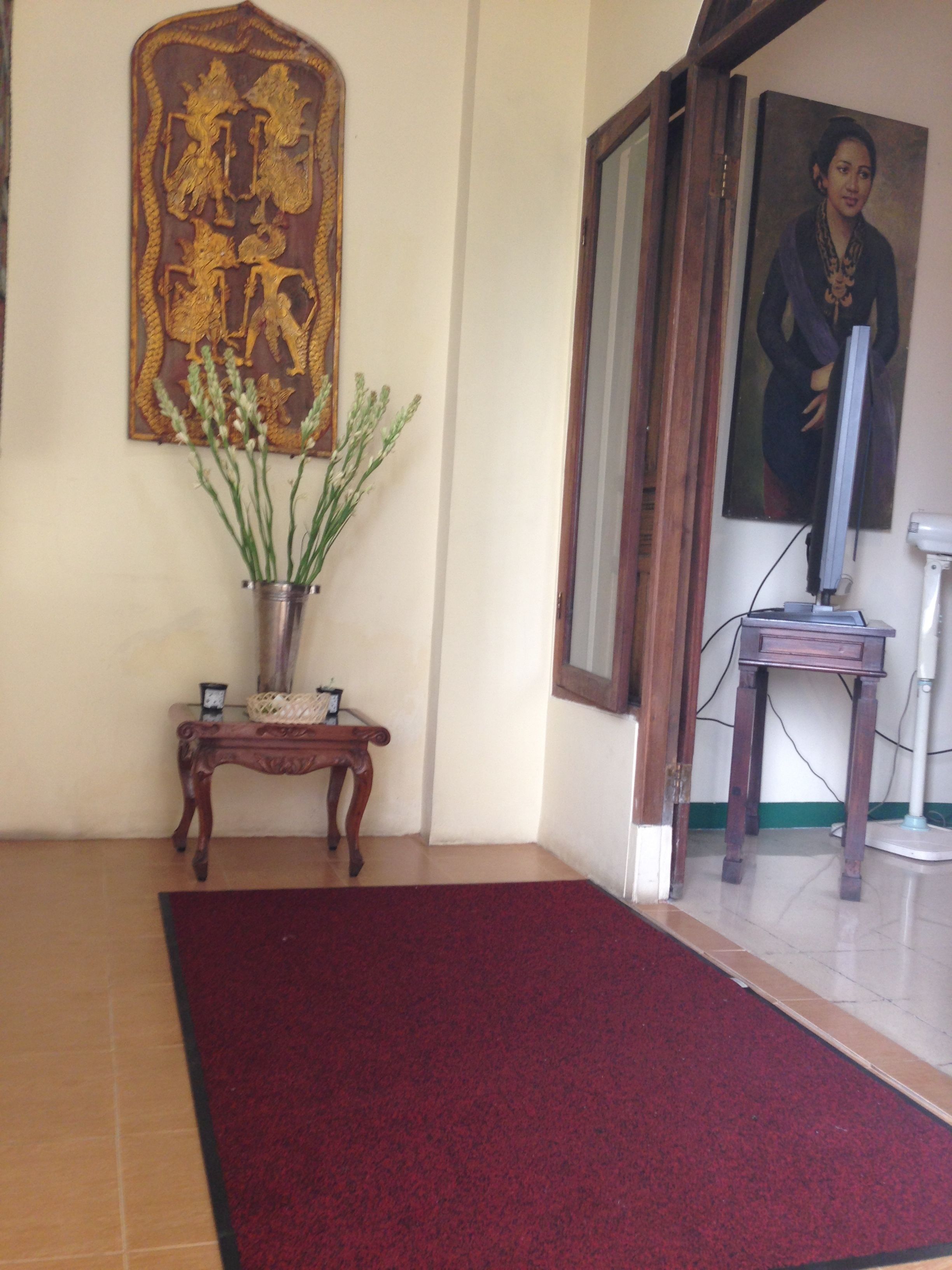 Quarto com carpete vermelho, mesa consola de madeira com um vaso de plantas, um quadro na parede e uma televisão sobre um suporte.