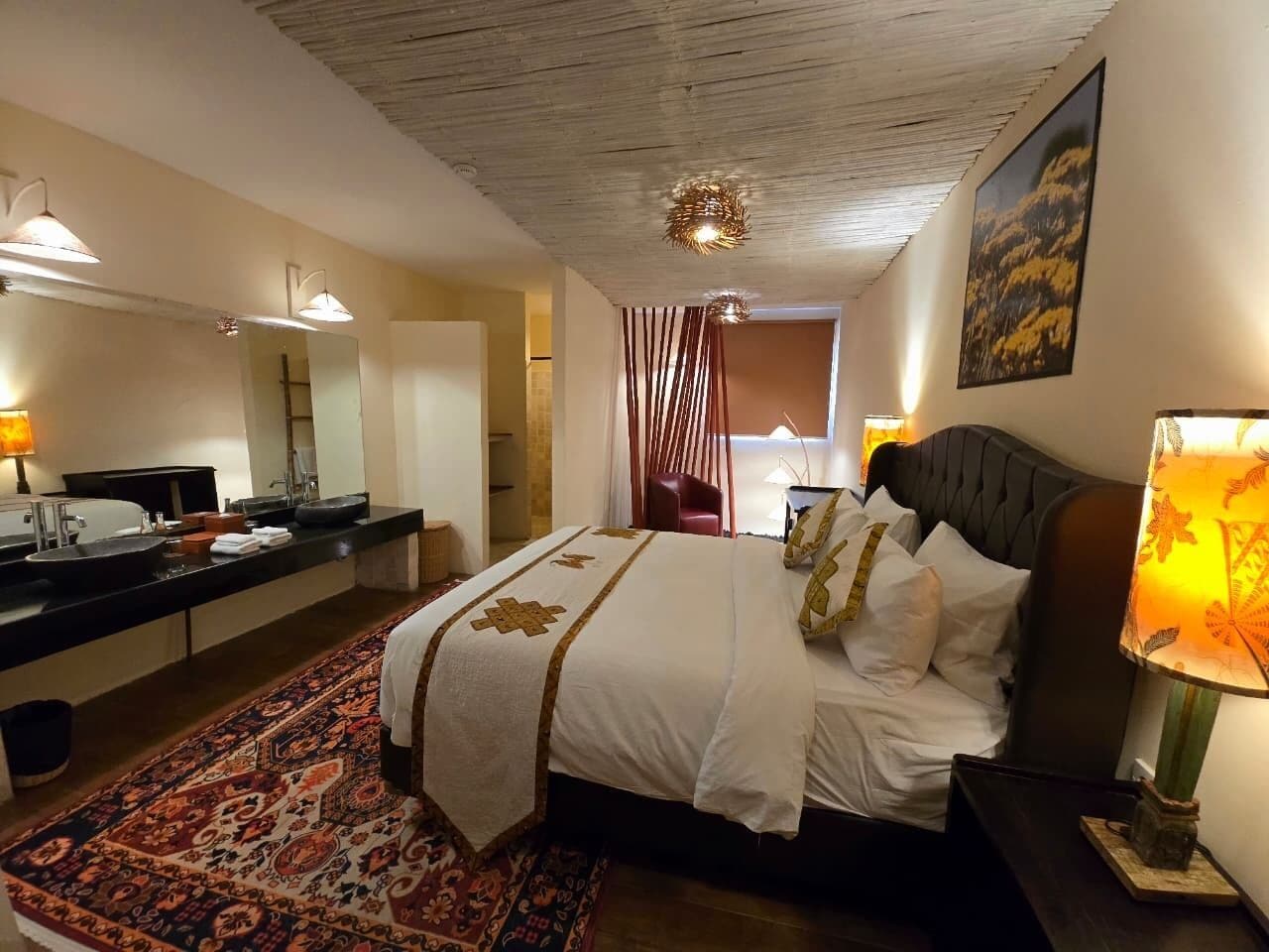 A hotel room with a large bed, a sitting area with a chair, a vanity with a mirror, and a window with curtains.