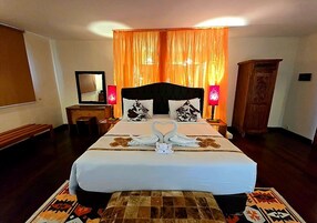 Uma cama bem arrumada com uma decoração de toalha em formato de coração, um banquinho de madeira, uma mesinha de cabeceira com abajur e um espelho na parede.