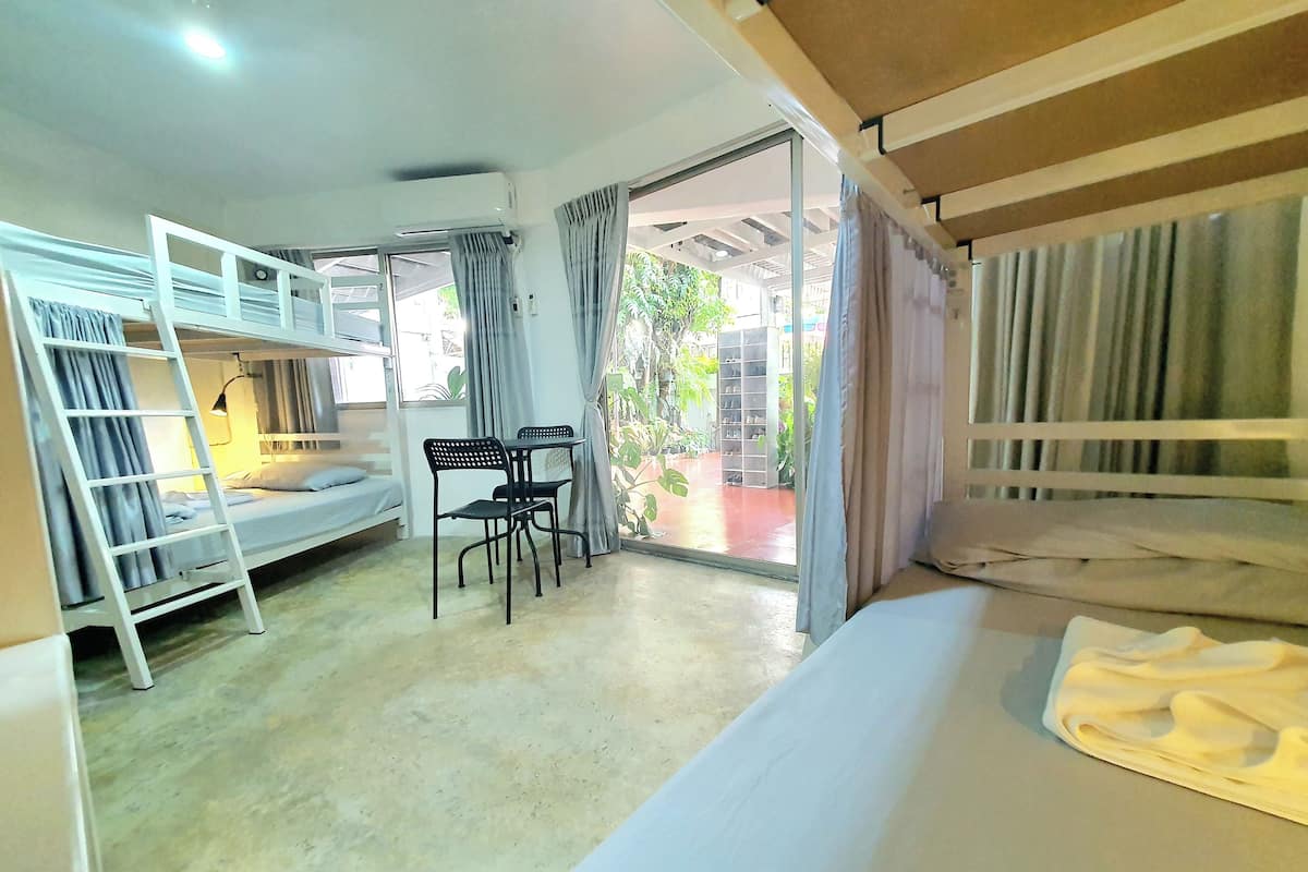 A dormitory room with bunk beds, a desk, chairs, and a window with curtains.