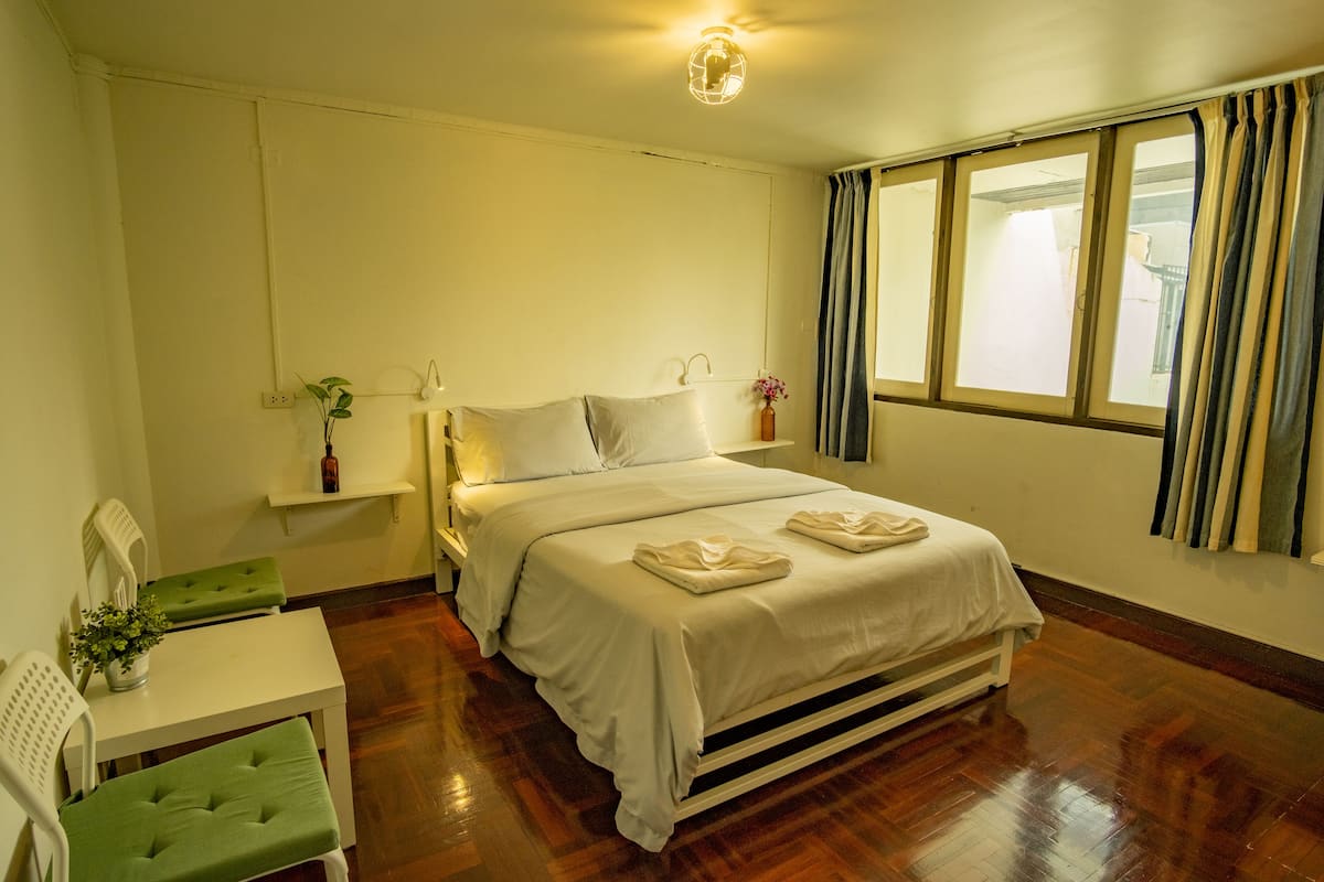 A hotel room with a bed, a green chair, a small table, a plant, and a window with curtains.