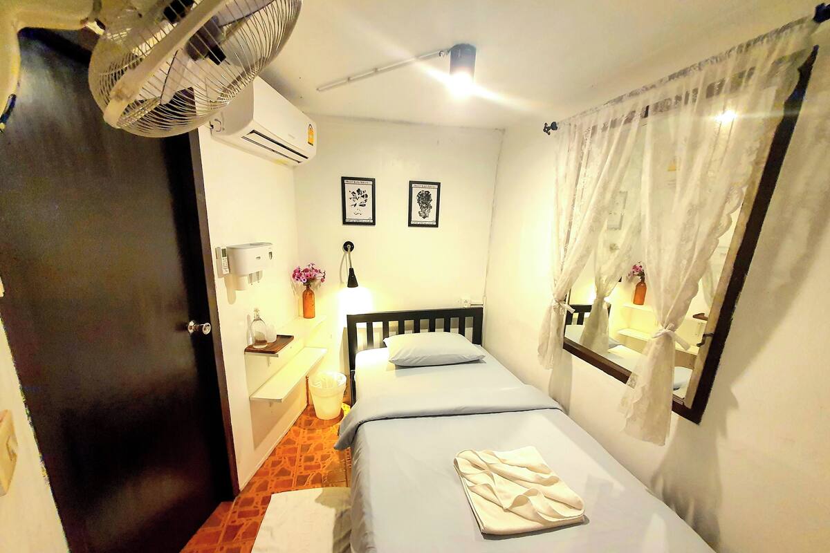 A hotel room with a bed, a bedside table, a lamp, a mirror, and an air conditioning unit.