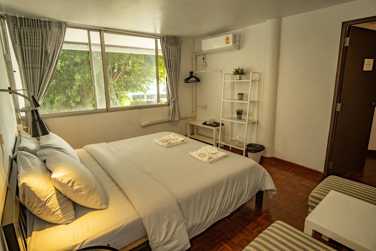 A neatly arranged hotel room with a large bed, a bedside table, a lamp, a small shelf with plants, and a window with curtains.