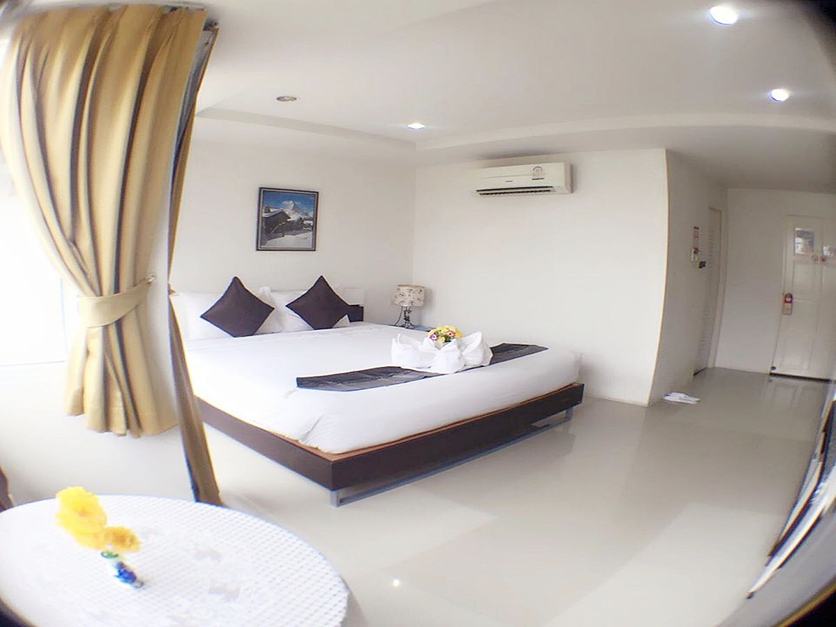 A hotel room with a bed, a bedside table with a lamp, an air conditioning unit, and a door.