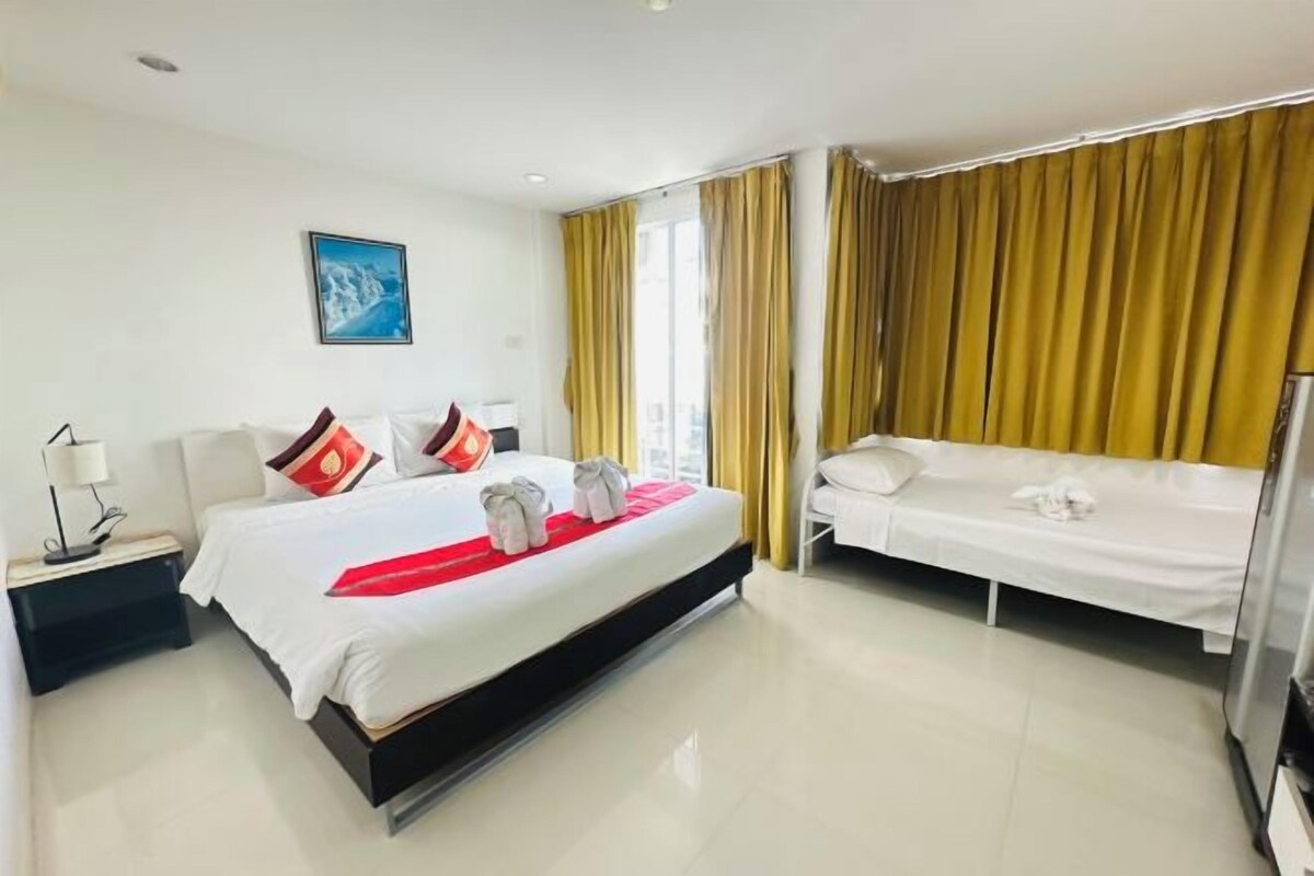 Quarto de hotel com uma cama grande, uma cama menor, uma mesa de cabeceira com abajur e uma janela com cortinas.