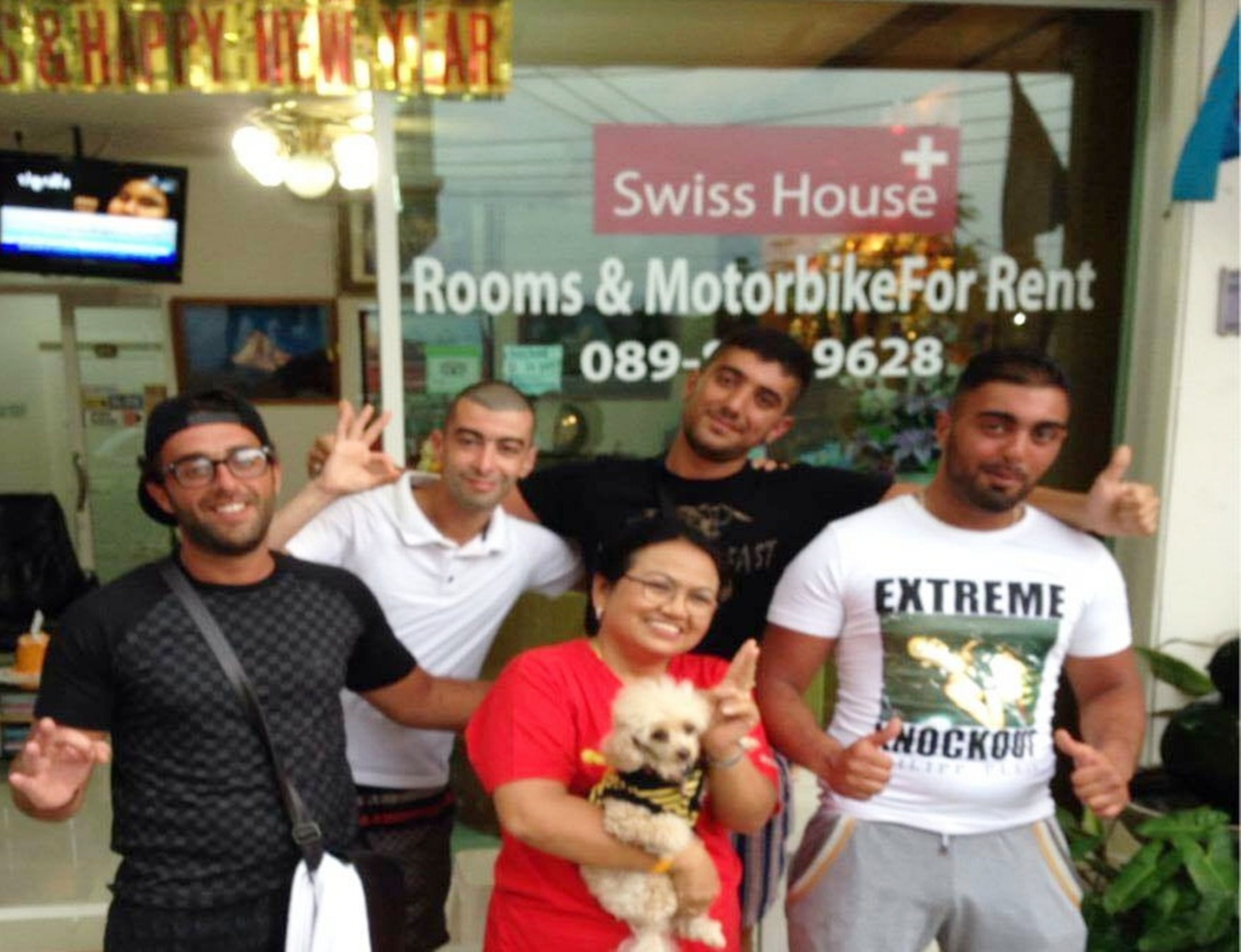 A group of people posing in front of a window with a sign for Swiss House, Rooms & Motorbike For Rent, and a phone number.