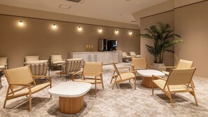Un vestíbulo de hotel con mostrador de recepción, zona de estar con sillas y mesas, y una planta grande en maceta.