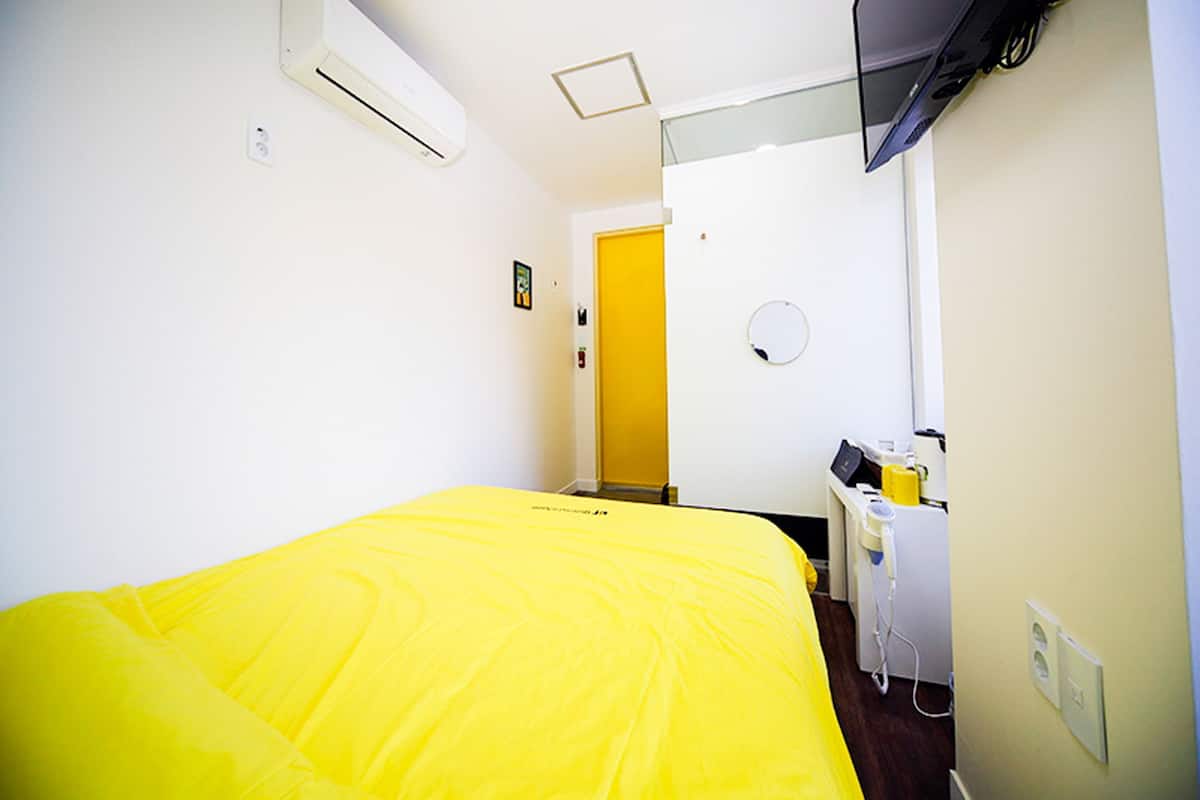 Ein Hotelzimmer mit Bett, Klimaanlage, Fernseher, einem kleinen Schreibtisch mit Stuhl und einer Tür zu einem anderen Raum.