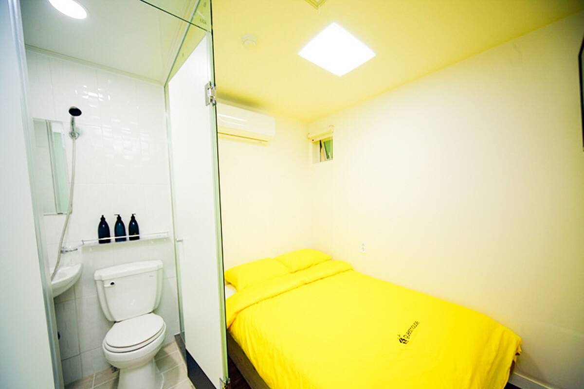 Ein Badezimmer mit Toilette, Waschbecken und Dusche. Ein Bett mit gelber Bettdecke ist vorhanden.