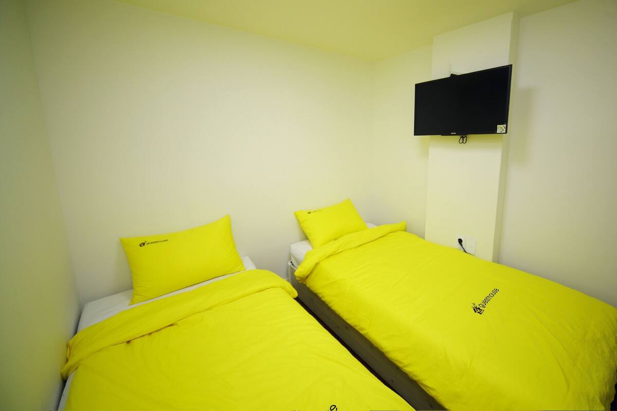 Ein Zimmer mit zwei Betten, jeweils mit einer hellgelben Tagesdecke und passenden Kissen, einem an der Wand befestigten Flachbildfernseher und einer Deckenleuchte.
