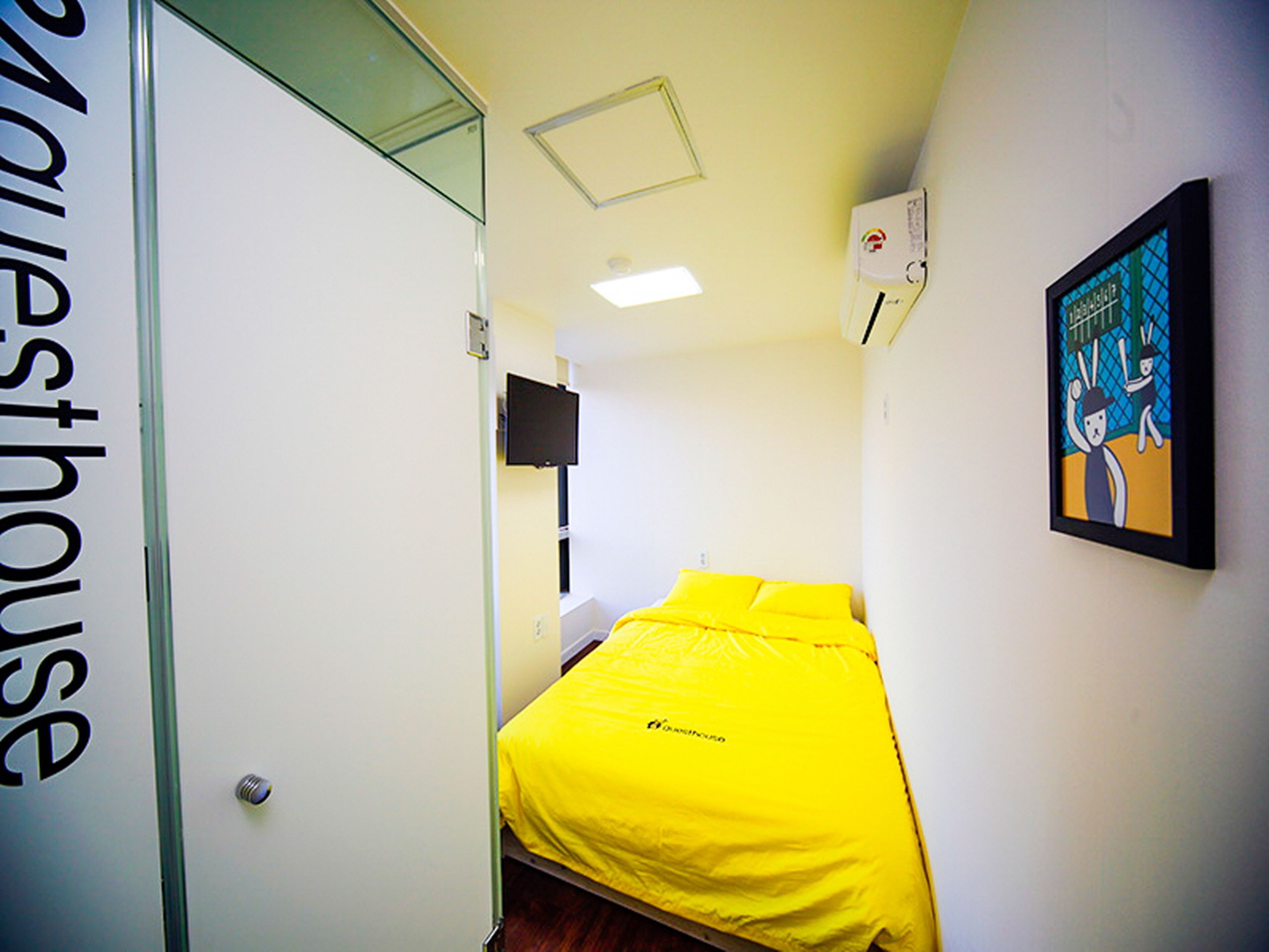 Een kleine, helder verlichte kamer met een bed met gele beddengoed, een flatscreen-televisie aan de muur en een kunstwerk met een tekenfilmfiguur.