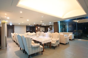 A spacious dining area with a long table and multiple upholstered chairs, a clock on the wall, and a large window offering a view of the outdoors.