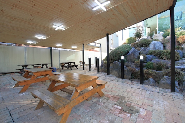 An outdoor patio area with wooden picnic tables, a brick floor, and a view of a landscaped garden with rocks and shrubs.