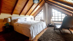 A bedroom with a wooden ceiling, a bed with white linens, a chair, and a window with curtains.