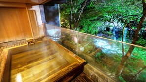 Un baño de aguas termales de madera con cerramiento de vidrio que da a una zona boscosa con cascada.