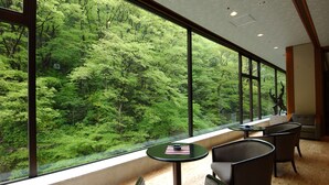 Una habitación con grandes ventanales que ofrecen vistas a un bosque frondoso. Hay dos sillas y una mesita redonda con un libro y una taza.
