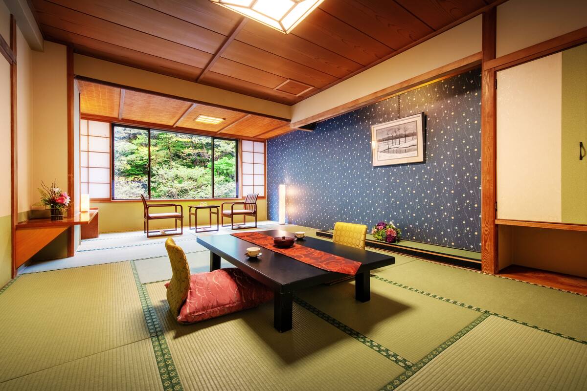 A traditional Japanese room with tatami flooring, a low wooden table, and a view of greenery through large windows.