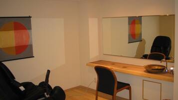 A room with a wooden desk, a chair, a guitar, a mirror, and a wall with a geometric painting.