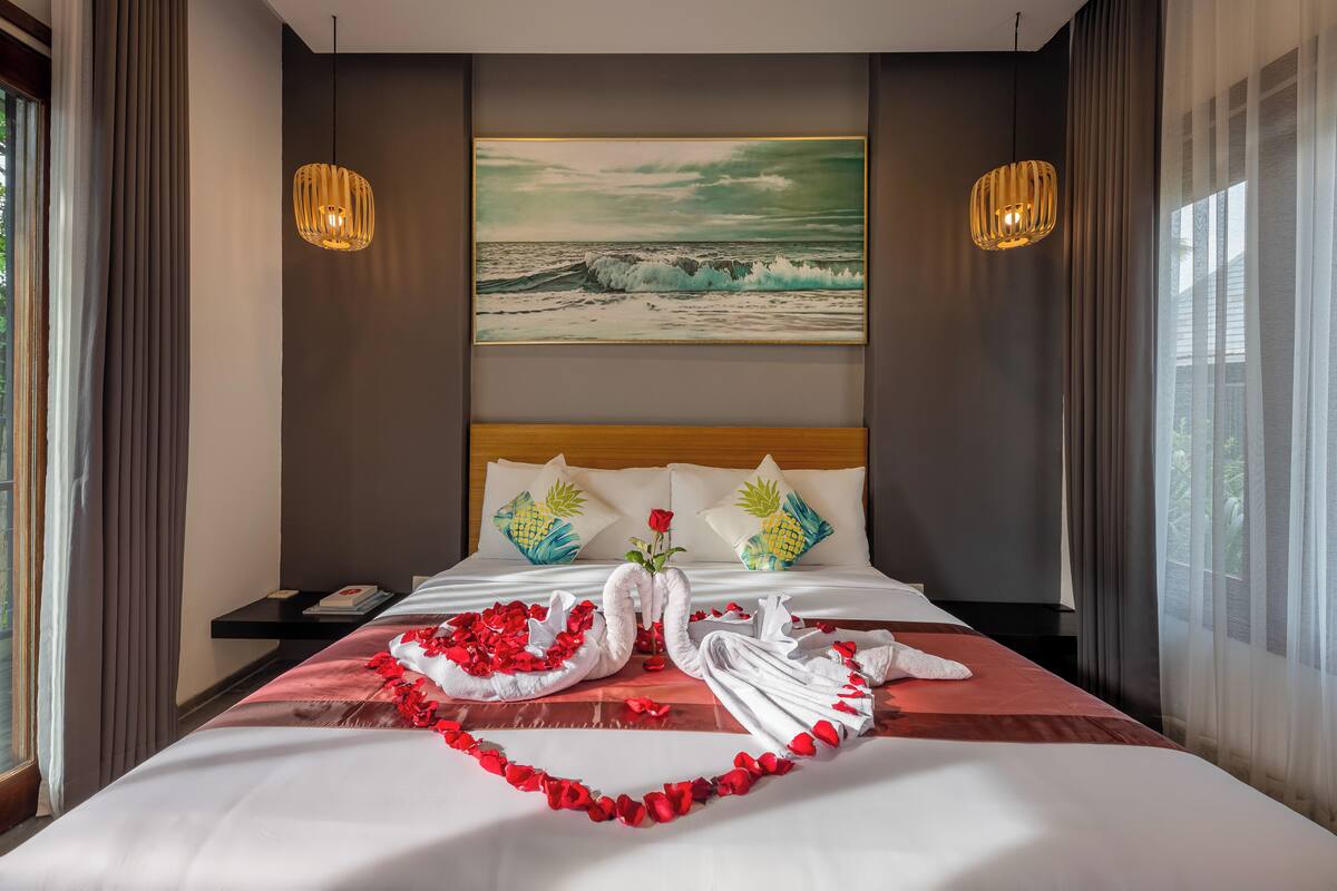 A hotel room with a large bed, a painting of the ocean, two wall-mounted lamps, and a nightstand with a book and a remote control.