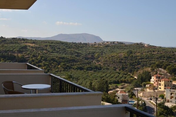 A balcony with a table and chair, overlooking a hillside with buildings and a mountain in the distance.