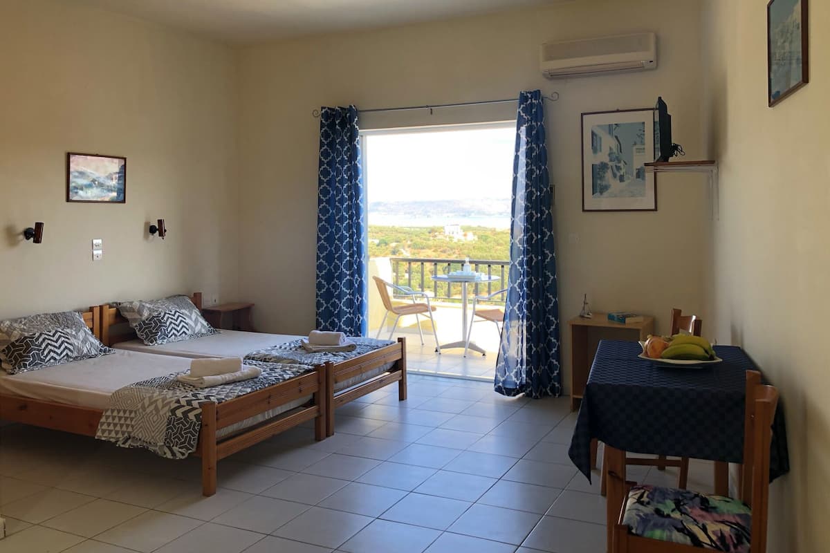 A room with two wooden beds, a small table with a plate of fruit, a chair, and a balcony with a view.