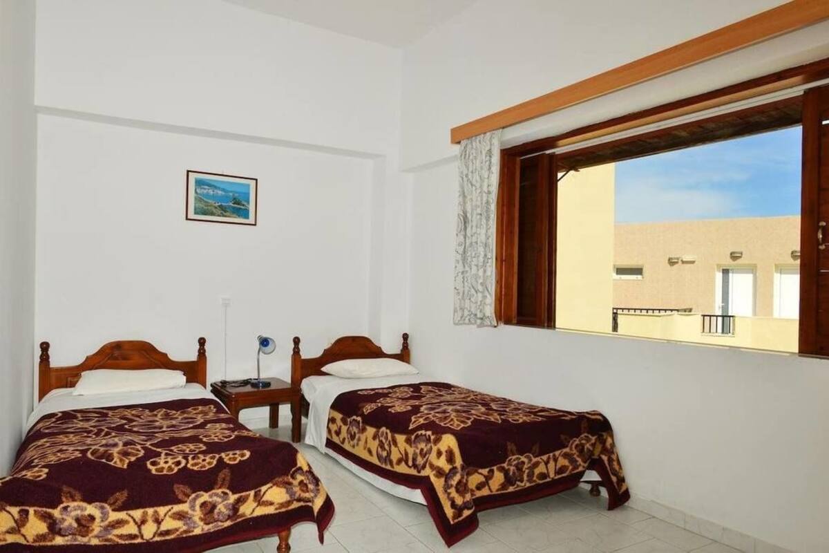 A room with two wooden beds, a small table, a lamp, and a window with a view of a building outside.
