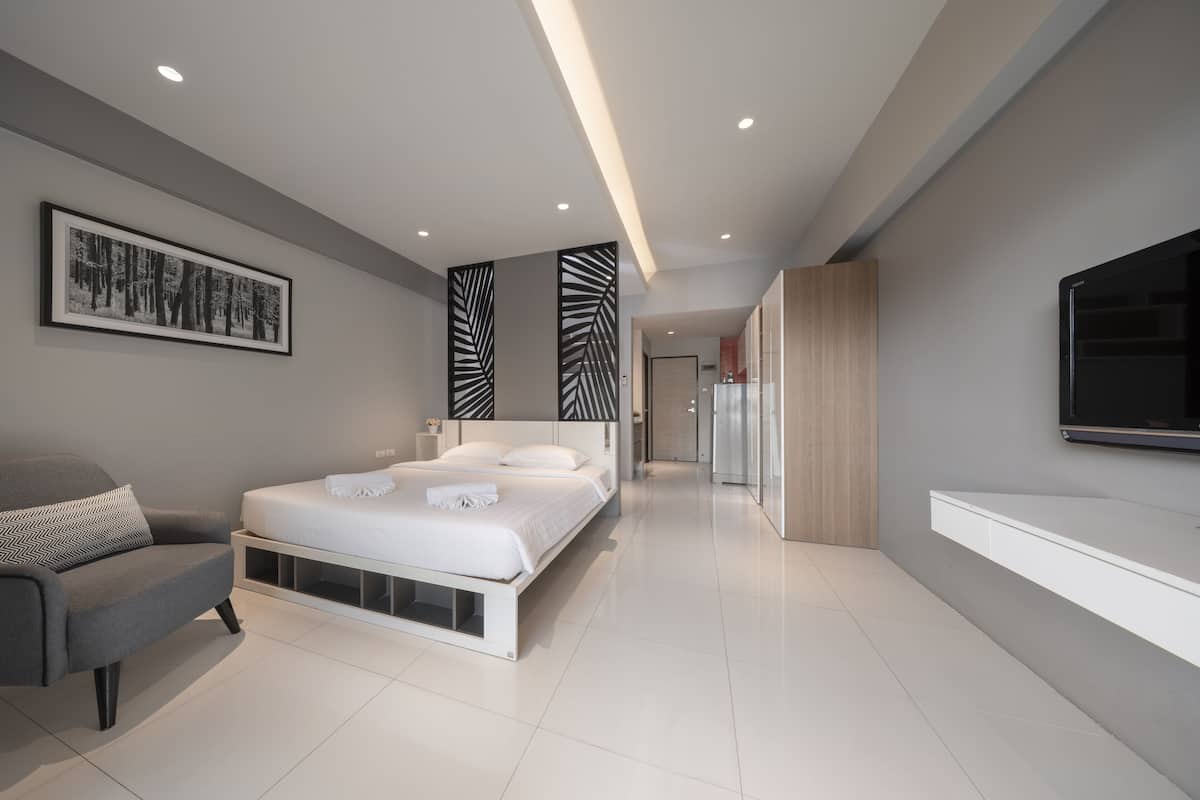 A modern bedroom with a bed, a chair, a TV, and a hallway leading to a kitchen.
