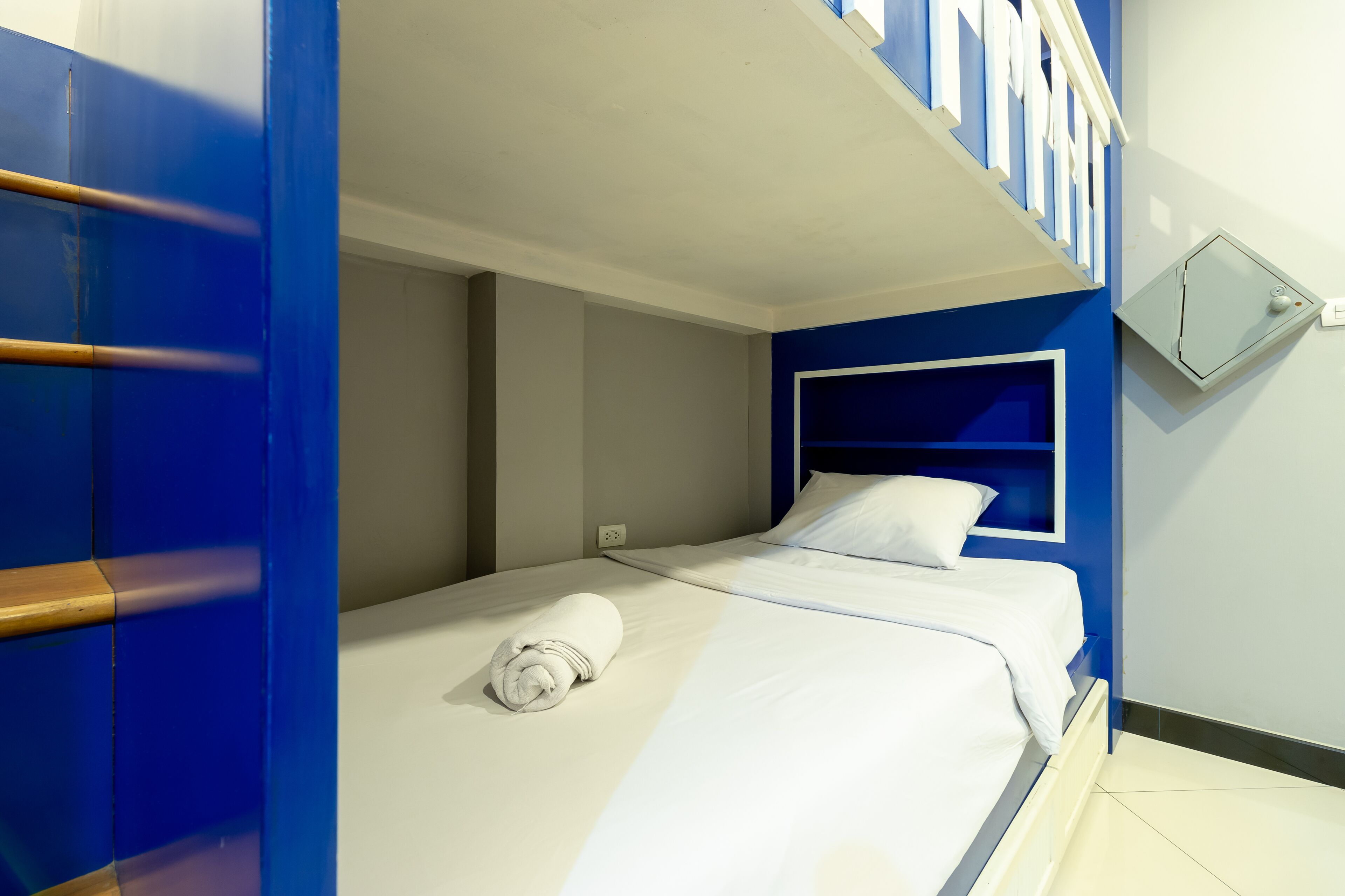 A single bed with a white mattress and a rolled white towel, a blue headboard, and a small bedside table with a white lamp.