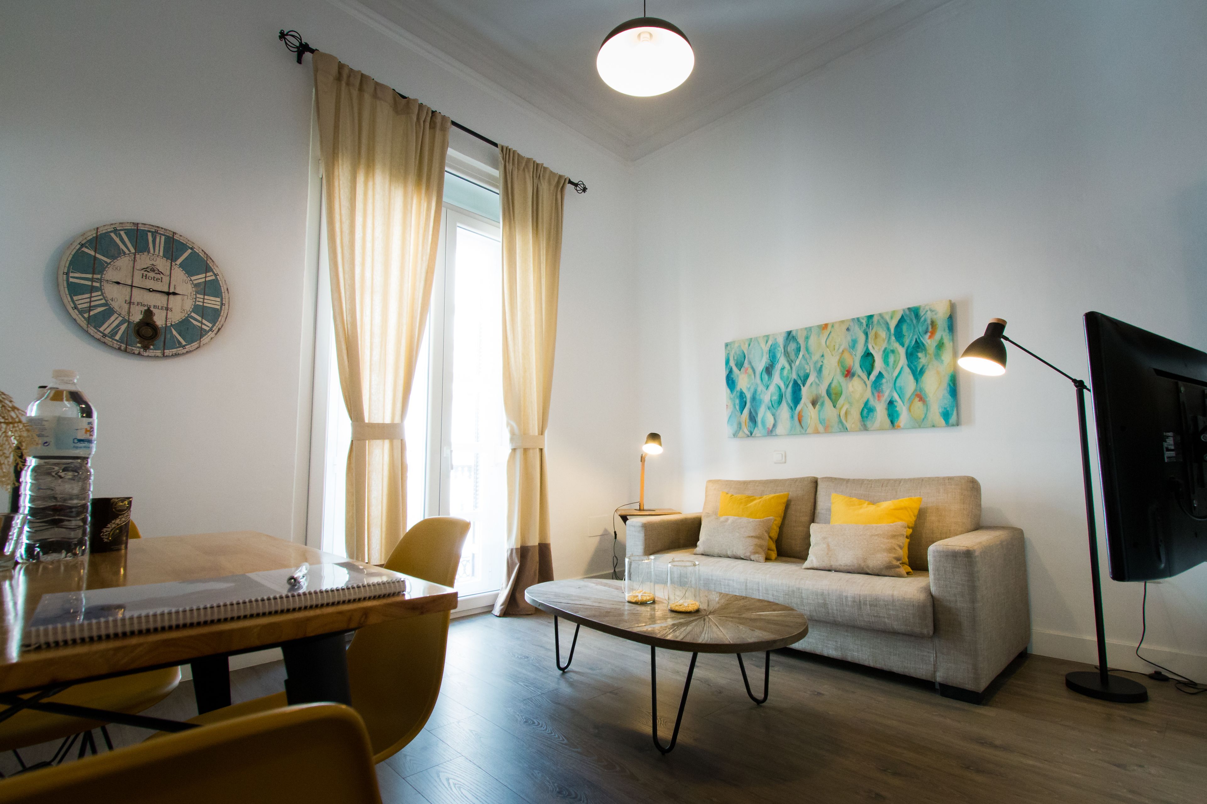 Uma sala de estar com sofá, mesa de centro, mesa de jantar com cadeiras, relógio, abajur e televisão.