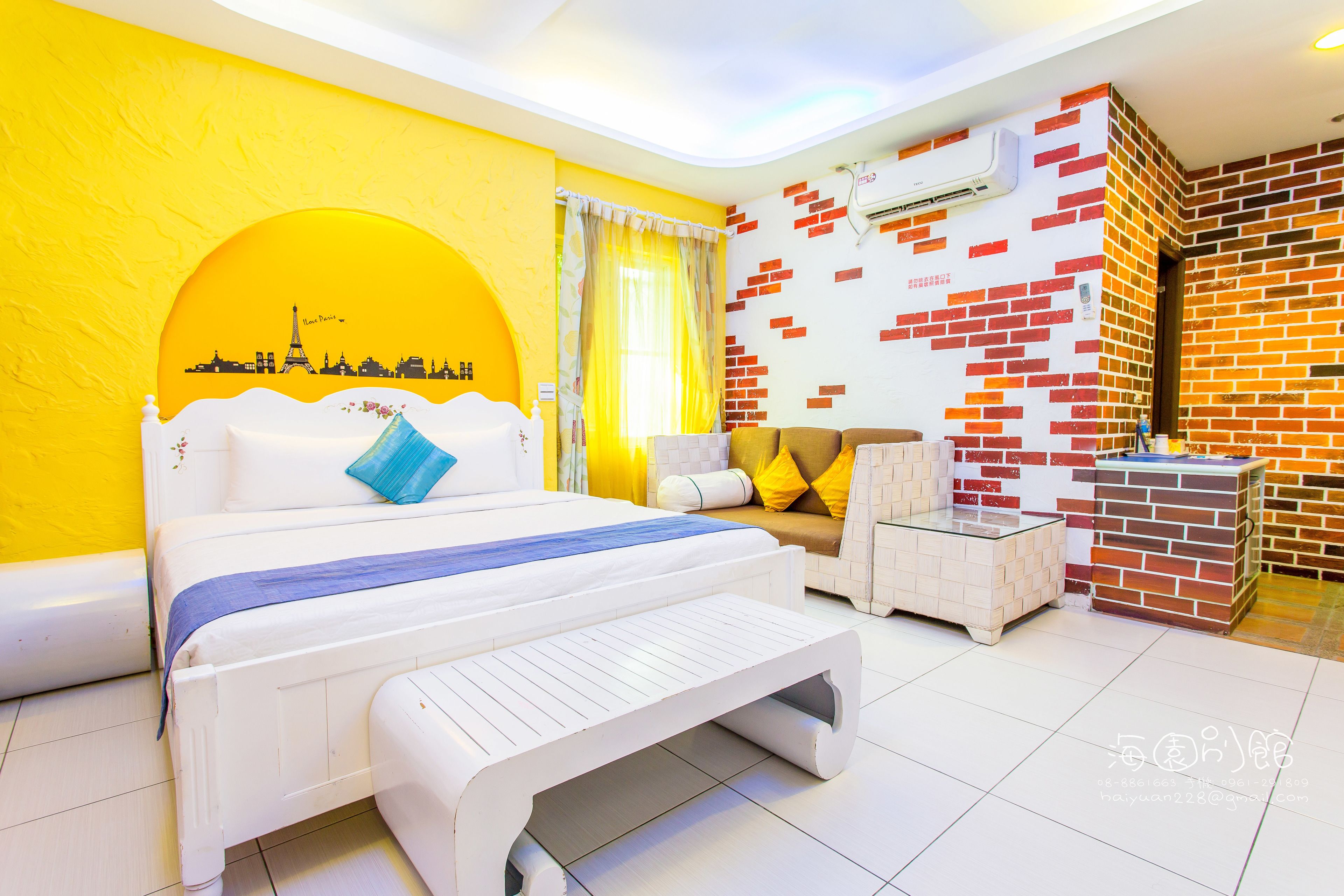 A brightly colored hotel room with a bed, a sofa, a small table, and a brick-patterned wall.