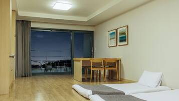 A hotel room with a wooden floor, a bed with white bedding, a desk with two chairs, and a balcony with a view of the city.