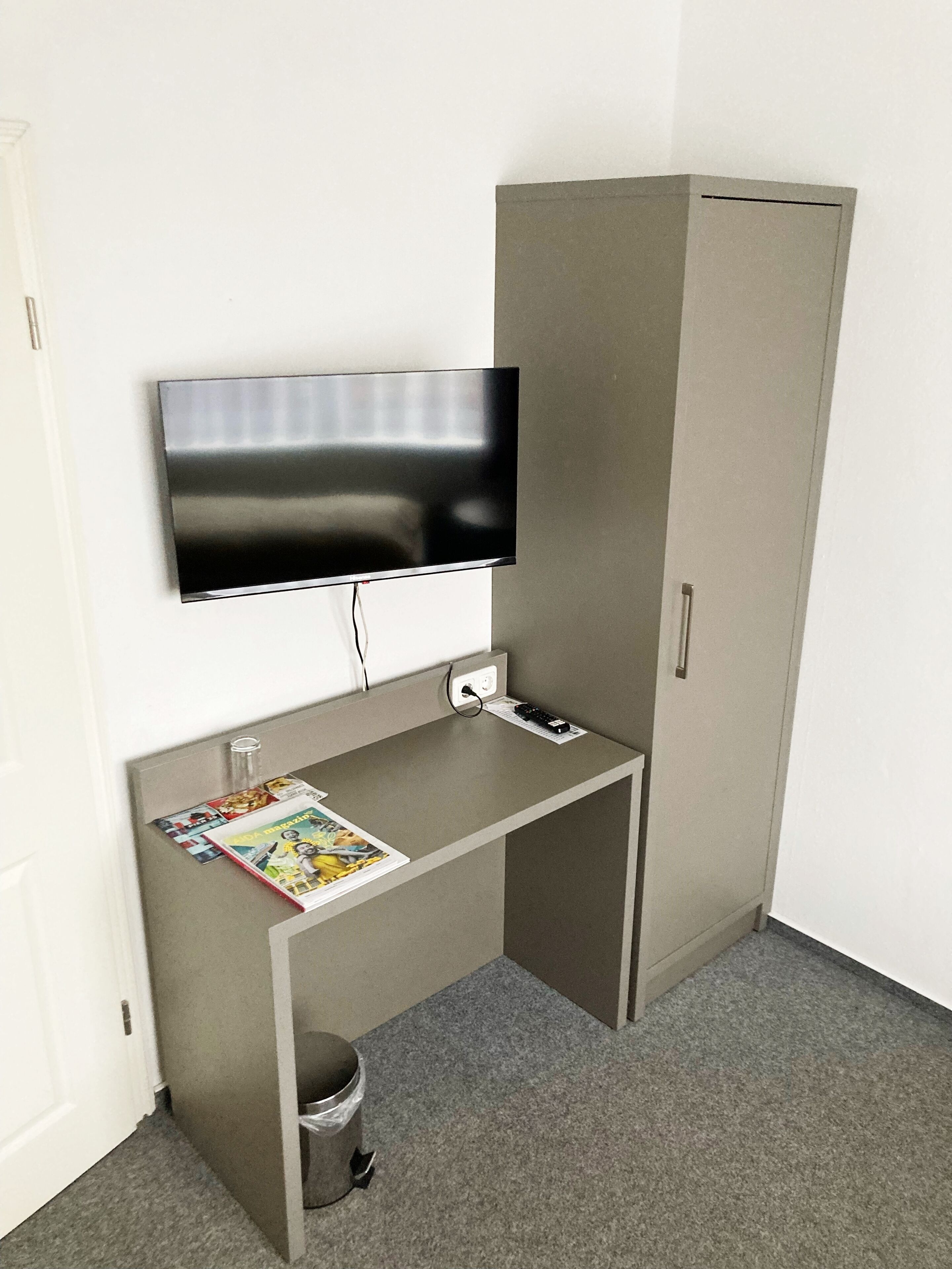 Une petite pièce moderne, avec un meuble gris, un téléviseur à écran plat fixé au mur et un petit bureau sur lequel se trouve un magazine.