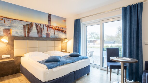 A hotel room with a bed, a desk, and a chair. There is a painting on the wall and a view of the outside through the window. - Familien- & Aparthotel Strandhof (Butjadingen)