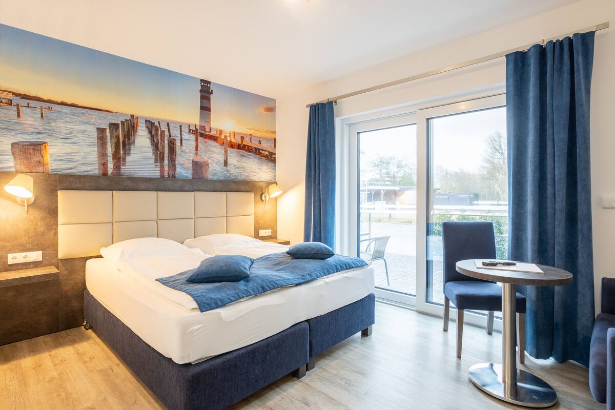 A hotel room with a bed, a desk, and a chair. There is a painting on the wall and a view of the outside through the window.