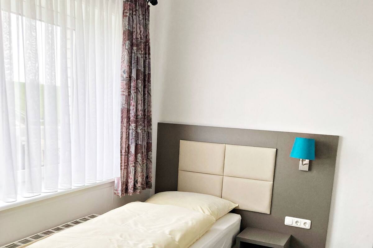 A hotel room with a bed, a small bedside table, a lamp, a chair, and a window with curtains.