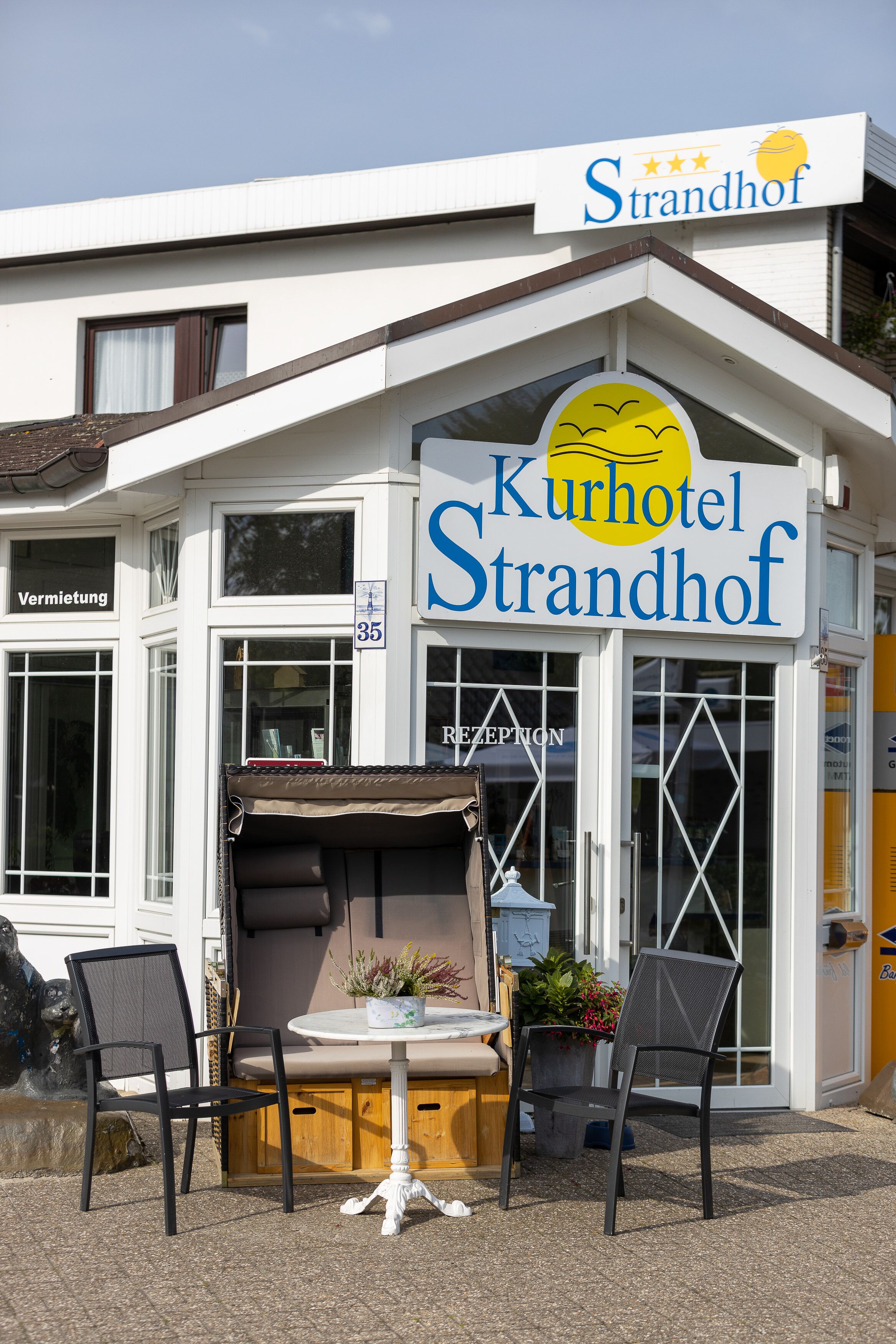 Hotel "Kurhotel Strandhof" met een bord waarop een zon en een vogel afgebeeld staan. Buiten staan twee stoelen en een kleine tafel.