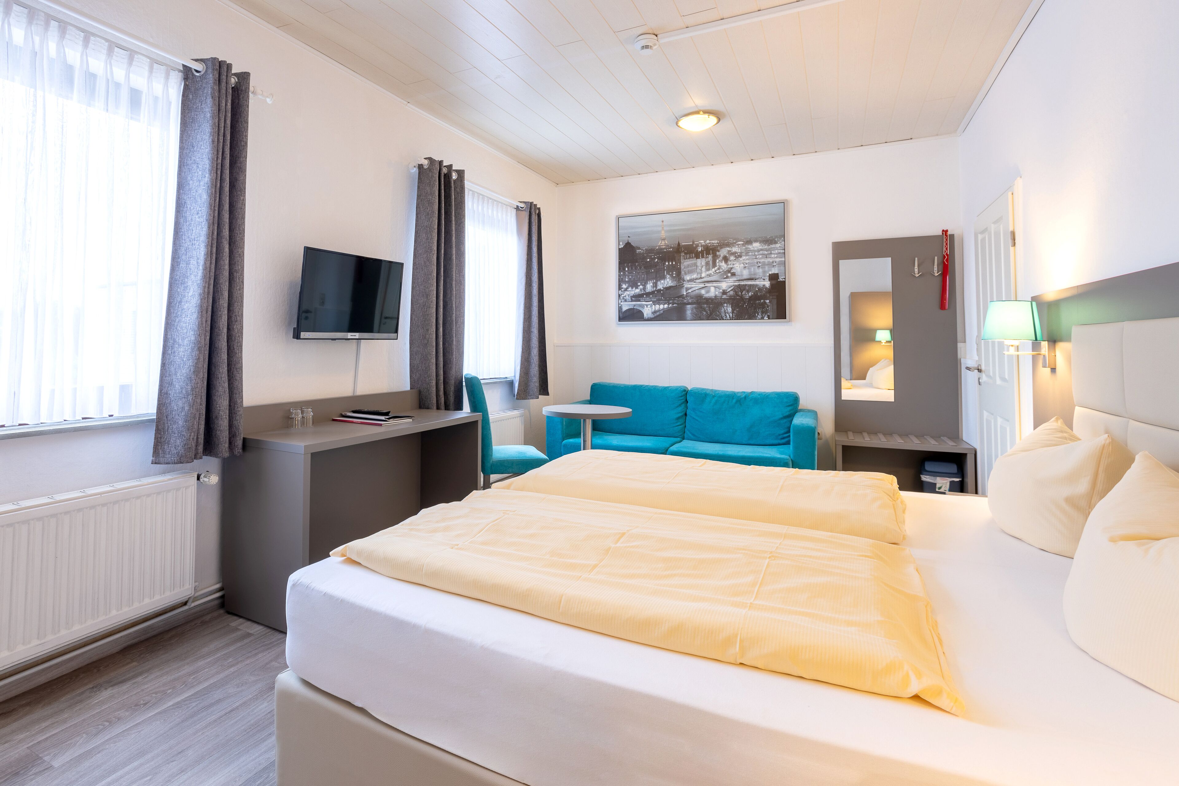 A hotel room with a bed, a desk, a chair, a small table, a TV, and a window with curtains.