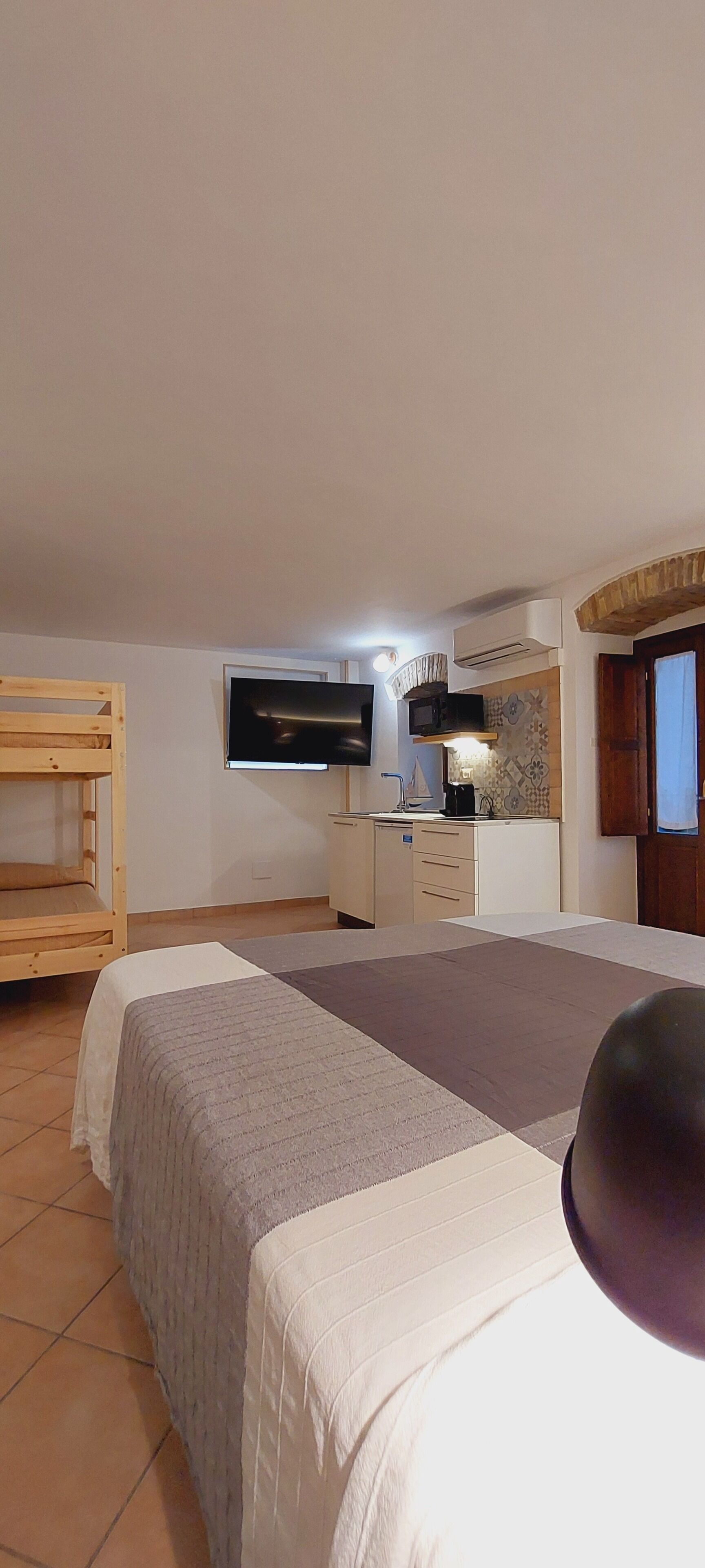 Un dormitorio con cama, literas de madera, kitchenette con televisor y aire acondicionado.