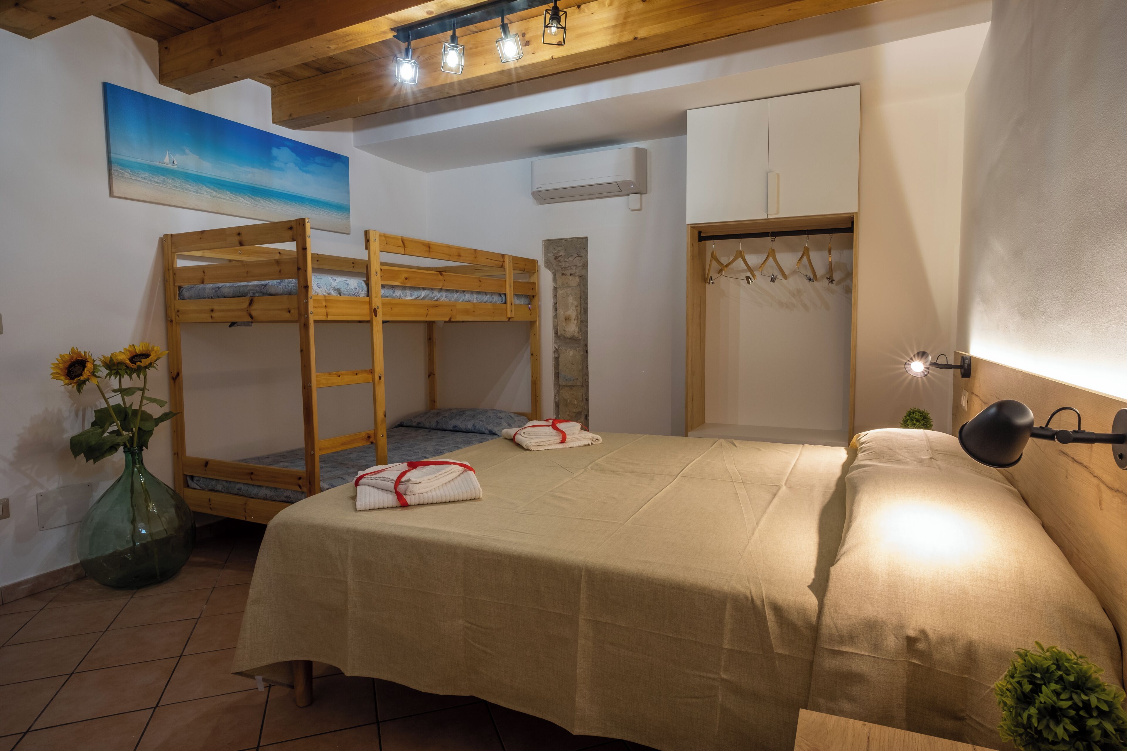 A bedroom with bunk beds, a bed with a beige cover, a wall-mounted wardrobe, a hanging coat rack, and a painting of a beach scene.