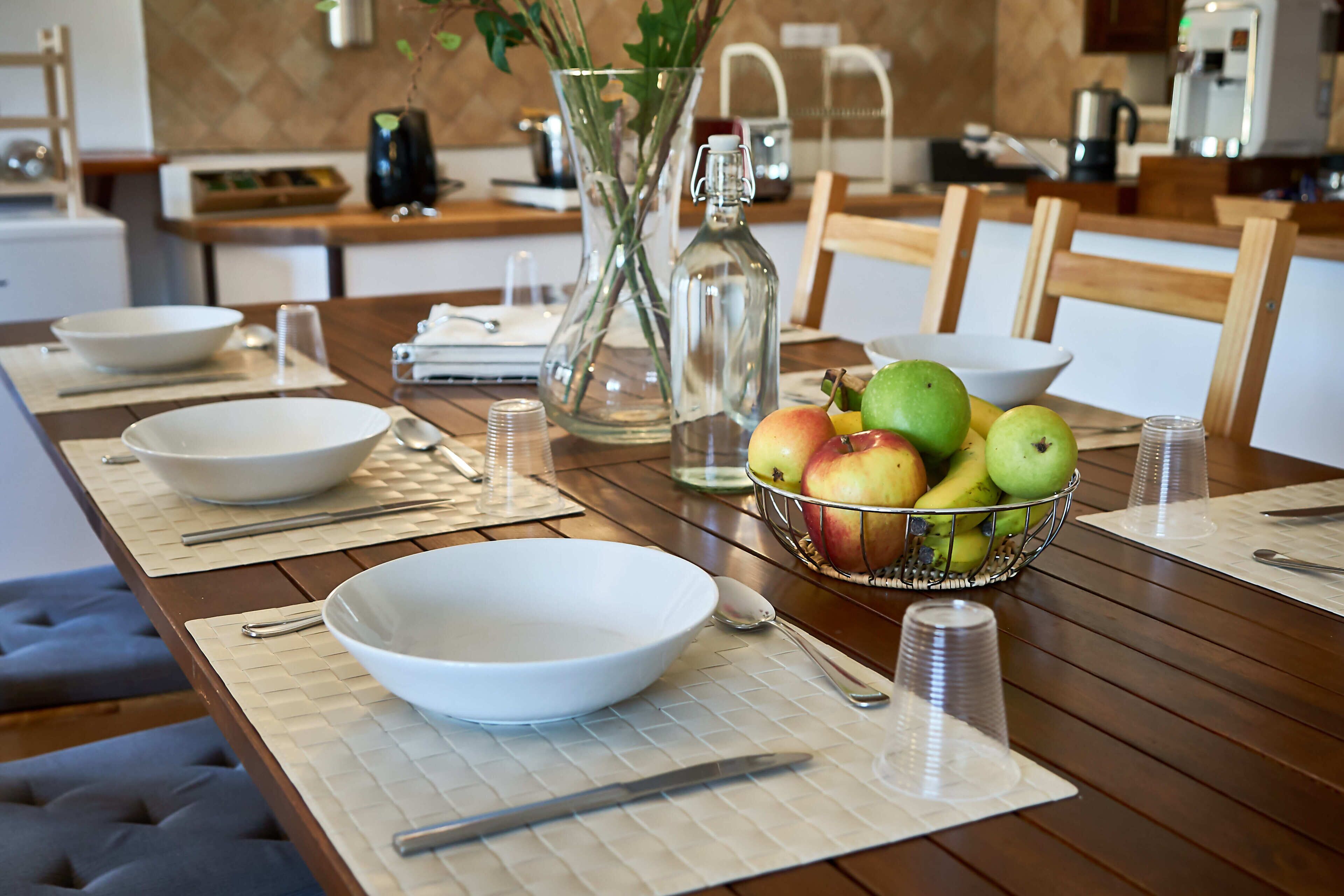 Une table à manger dressée avec des bols, des cuillères, un couteau, une bouteille en verre et un saladier rempli de fruits.