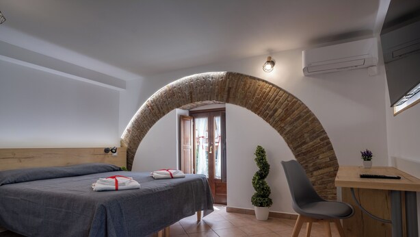 A bedroom with a bed, a desk, a chair, a potted plant, and an arched doorway. - B&B La Suite (Milazzo)