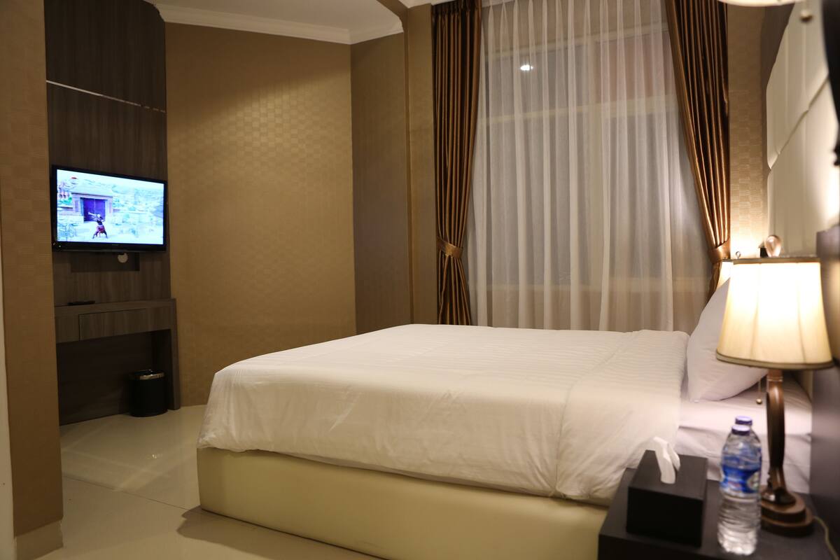 Una habitación de hotel con una cama, un televisor en la pared, una mesita de noche con una lámpara y una botella de agua, y cortinas.