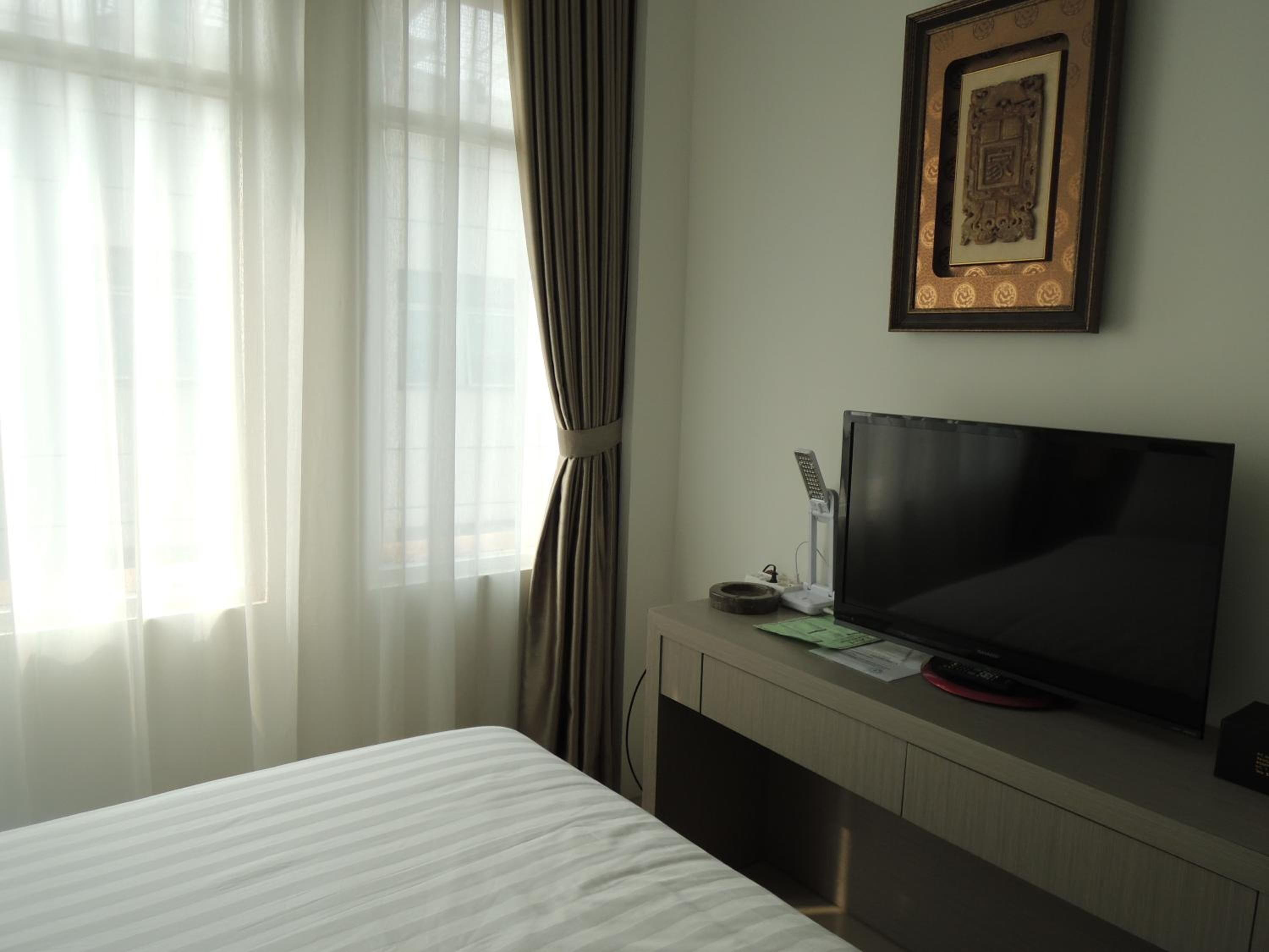 Habitación de hotel con cama, televisor sobre un soporte de madera, un cuadro en la pared y cortinas de encaje.
