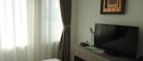 Habitación de hotel con cama, televisor sobre un soporte de madera, un cuadro en la pared y cortinas de encaje.