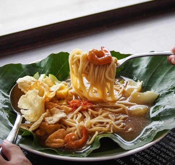 A bowl of noodles with shrimp, vegetables, and fried items on a leaf. - Hotel 55 (Jakarta)
