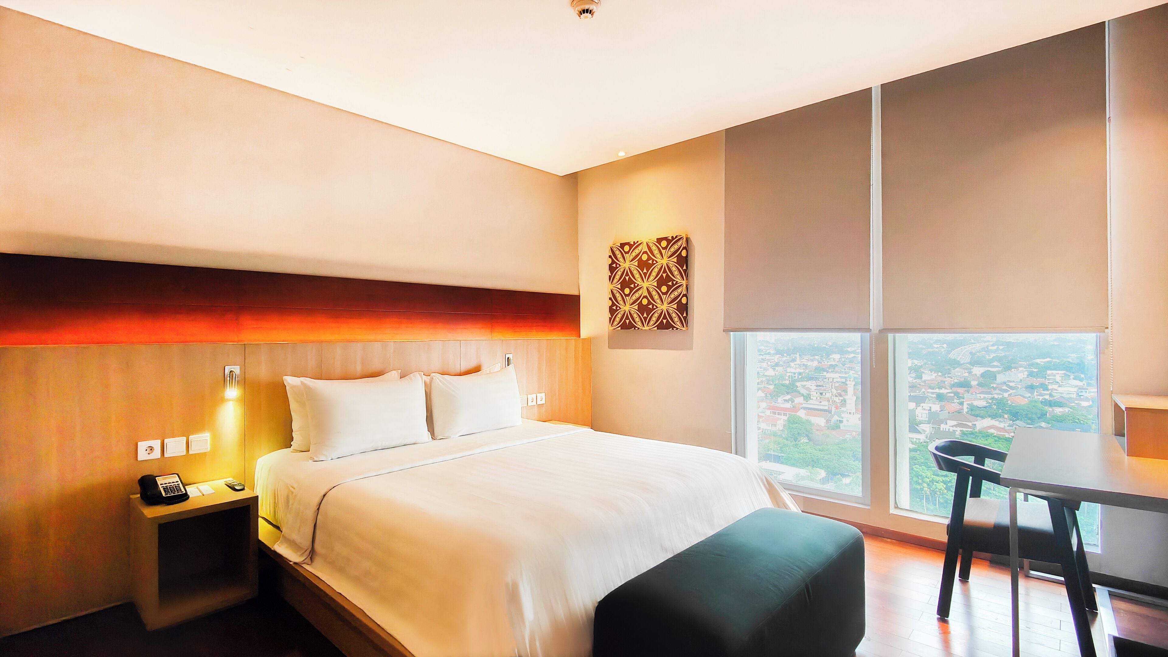 A modern hotel room with a large bed, bedside table with a phone, a desk with a chair, and a city view through large windows.