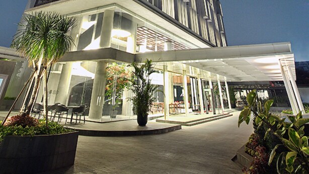 Un hotel moderno con una estructura alta y rectangular, un área de entrada cubierta con asientos y plantas en macetas.