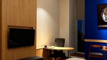 A hotel room with two beds, a desk with a chair, a flat-screen TV mounted on the wall, and a large window.