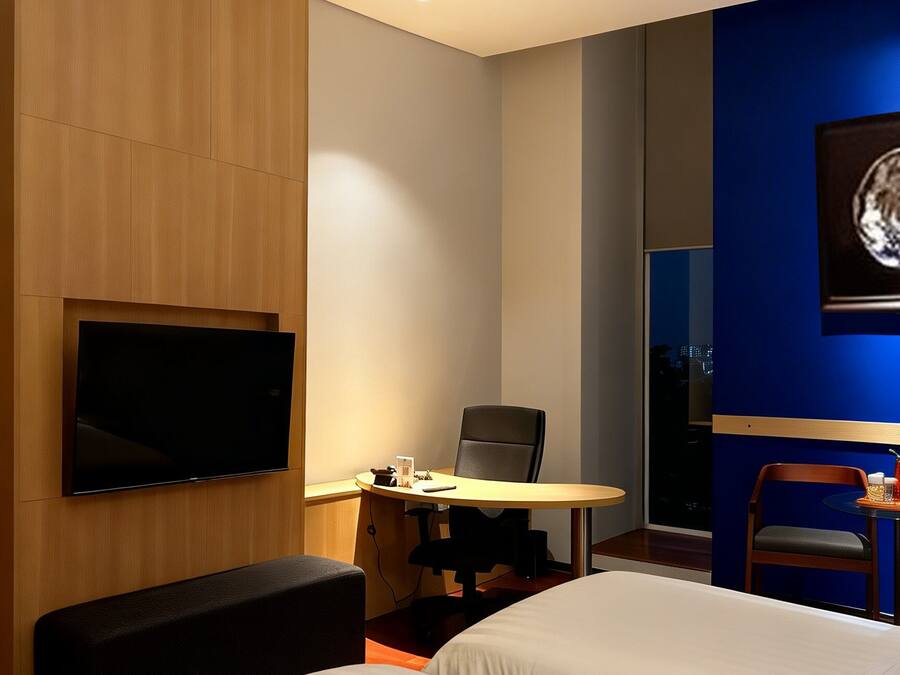 A hotel room with two beds, a desk with a chair, a flat-screen TV mounted on the wall, and a large window.