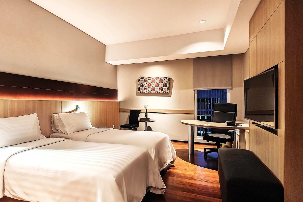 A hotel room with two beds, a desk, a chair, a TV, and a small table.
